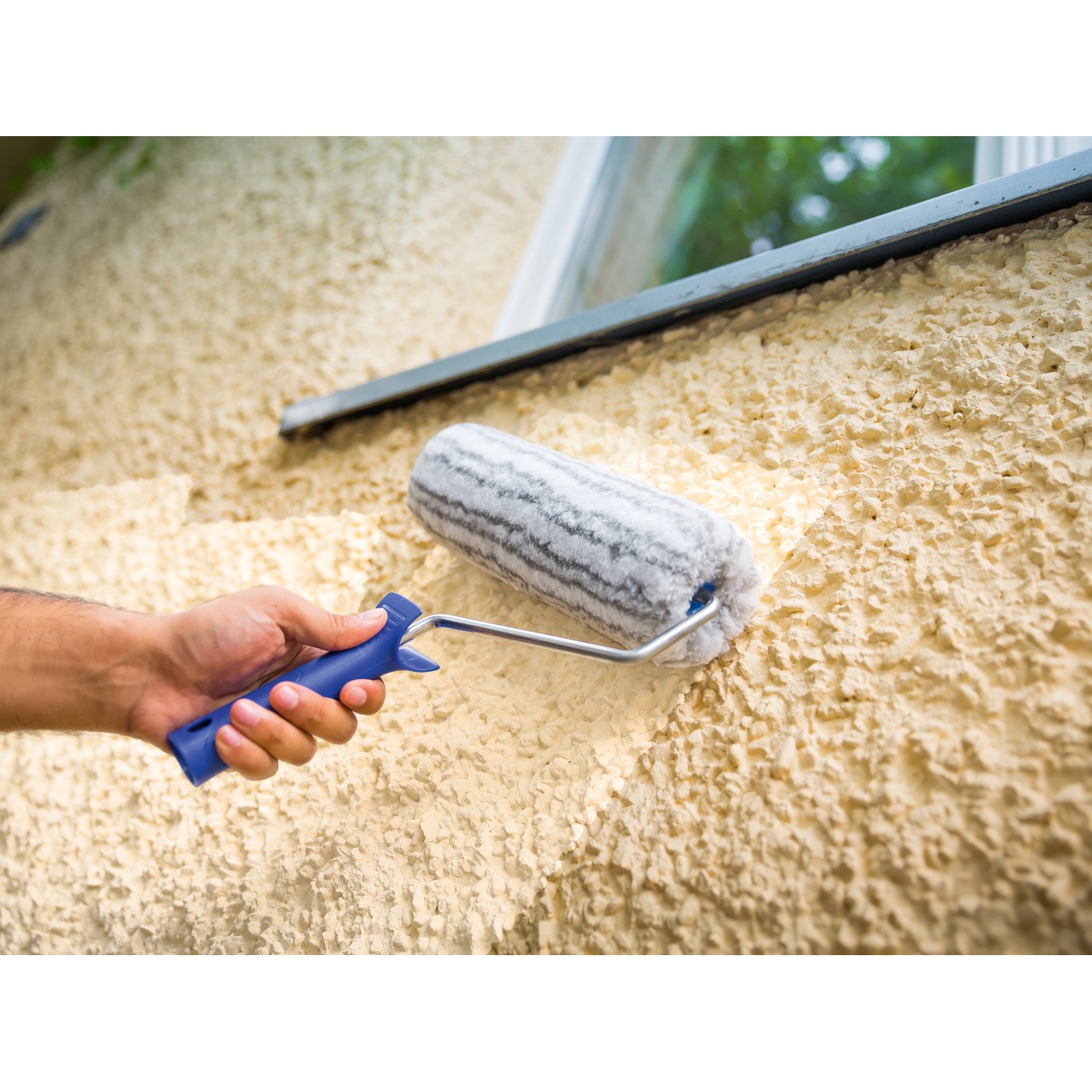 Hand streicht raue Wand mit LUX Dispersionswalze 25 cm. Farbwalze mit blauem Griff.