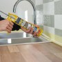 Person dichtet Küchenspüle mit transparentem Soudal Silikon ab. Gelbe Kartuschenpistole, Holzplatte, Fliesenwand.