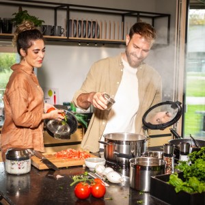 Paar kocht mit RÖSLE Silence Pro Edelstahl Topfset auf Induktionsherd. Mann würzt, Frau hält Pfanne. Frische Zutaten.