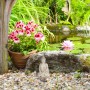 Cremeweiße Buddha Figur aus Polyresin, sitzend im Garten neben Teich und Blumen.