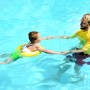 Kind mit gelbem Freds Swim Academy Schwimmreifen im Pool, unterstützt von einem Erwachsenen.