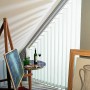 Weißer Lamellenvorhang gestreift im Dachfenster, mit Staffelei und Tisch im Vordergrund.