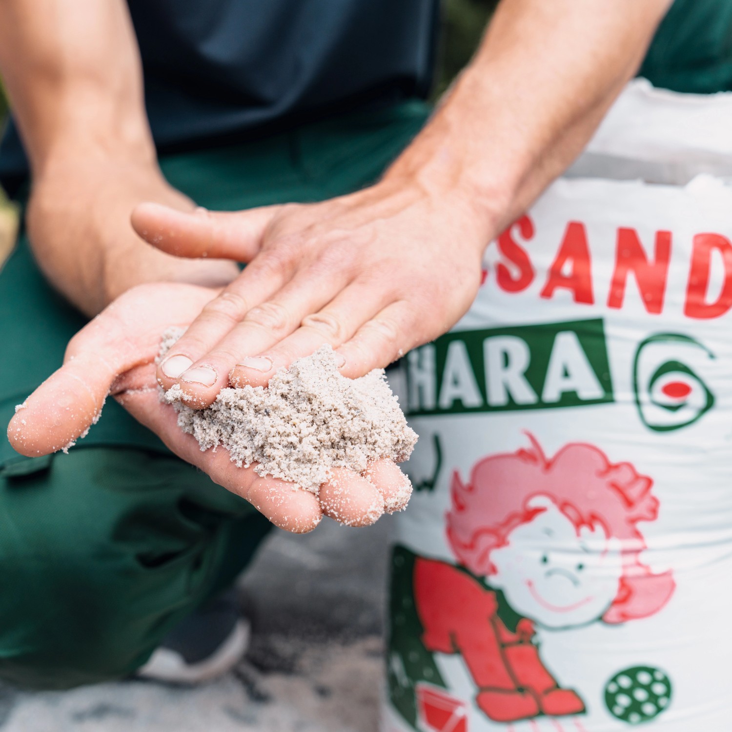 Weco Spielsand Sahara, 15kg Sack. Sand in der Hand vor Sack im Zierkies & Ziersplitt-Look.