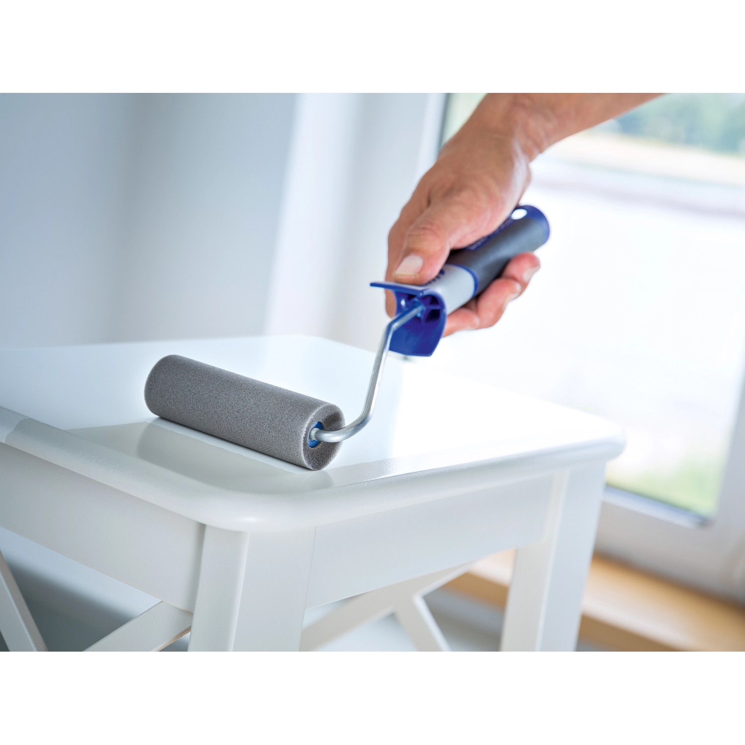 Hand hält LUX Schaumroller 11 cm beim Lackieren eines Tisches. Farbroller für glatte Untergründe.