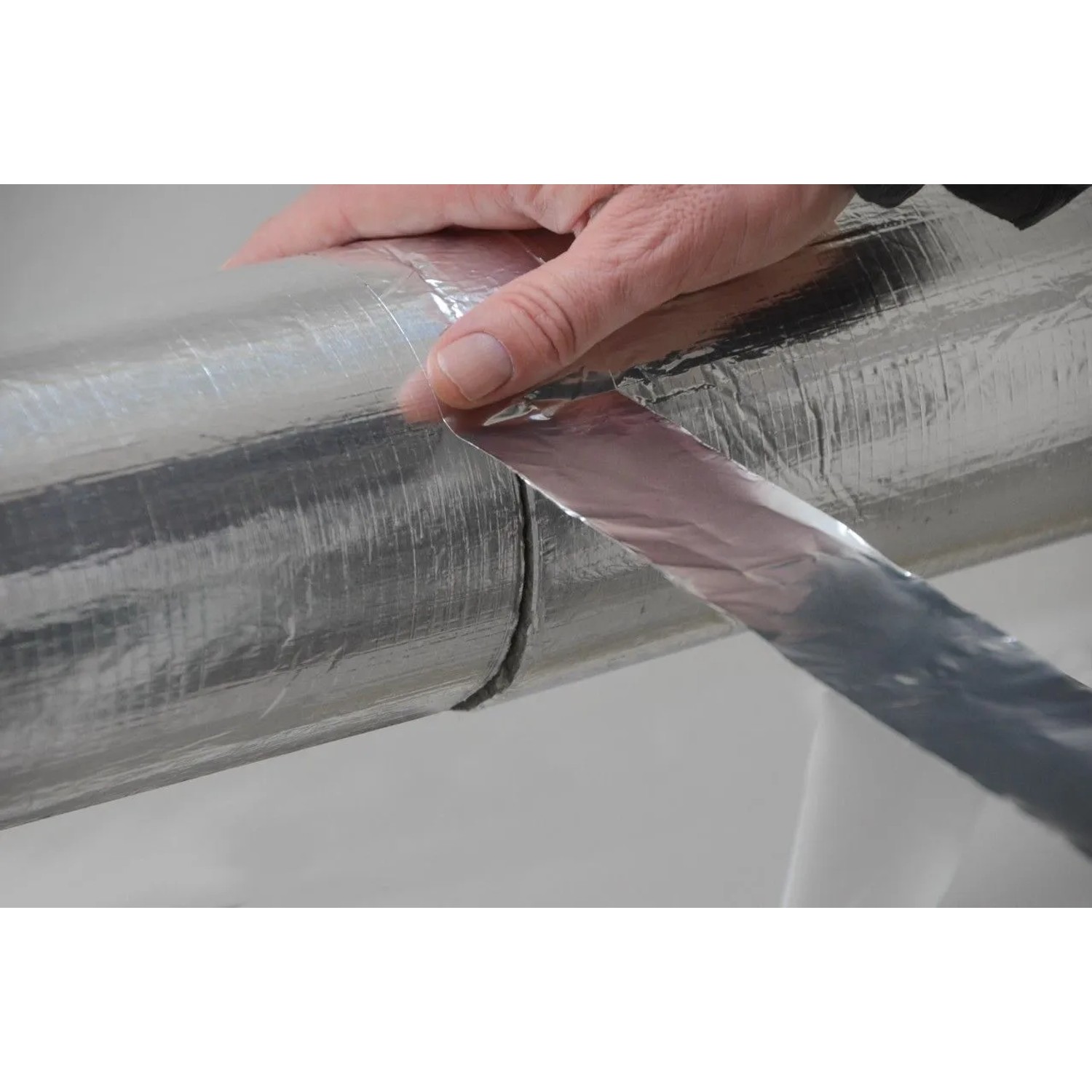 Hand klebt glänzendes Aluminium Klebeband auf eine silberne Rohrisolierung. Dämmstoffzubehör für Rohrschalen und Lamellenmatten.
