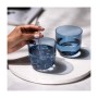 Zwei blaue Trinkgläser auf hellem Tablett. Eine Hand rührt Eis in einem Glas mit Strohhalm. Modernes Glas-Geschirr-Set.