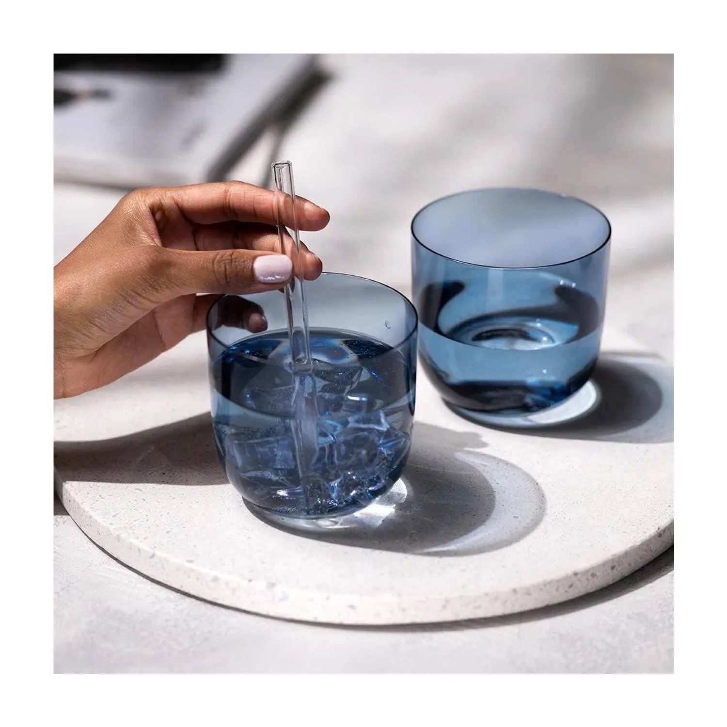 Zwei blaue Trinkgläser auf hellem Tablett. Eine Hand rührt Eis in einem Glas mit Strohhalm. Modernes Glas-Geschirr-Set.