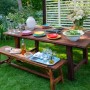 Gartenmöbel-Set aus Teakholz, behandelt mit Bondex Teak-Öl, mit Tisch, Bänken und farbenfrohem Geschirr.