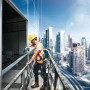 Arbeiter mit gelbem Helm und orangefarbener Weste bohrt mit Bosch Akku-Bohrschrauber an Hochhausfassade vor Stadtkulisse.