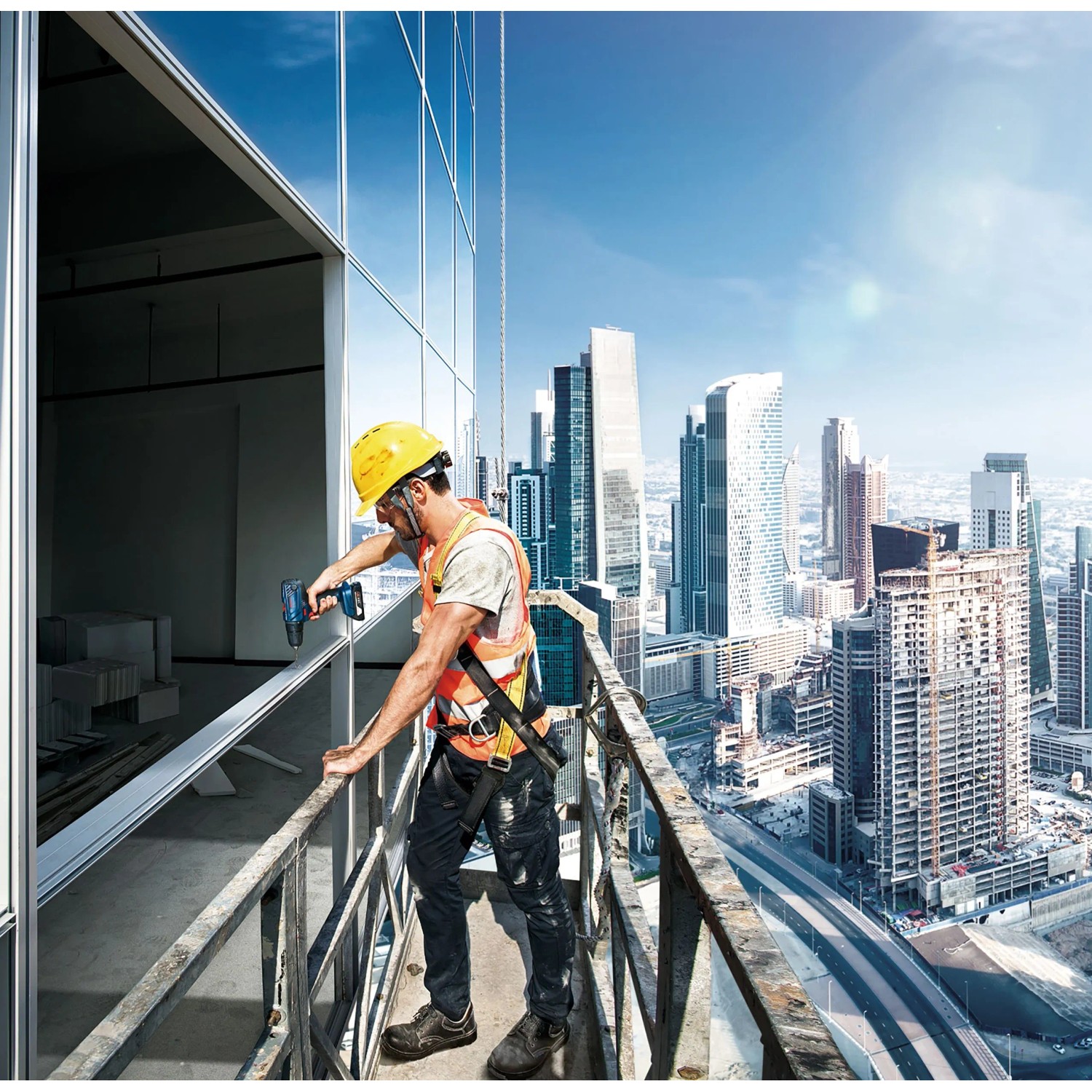 Arbeiter mit gelbem Helm und orangefarbener Weste bohrt mit Bosch Akku-Bohrschrauber an Hochhausfassade vor Stadtkulisse.
