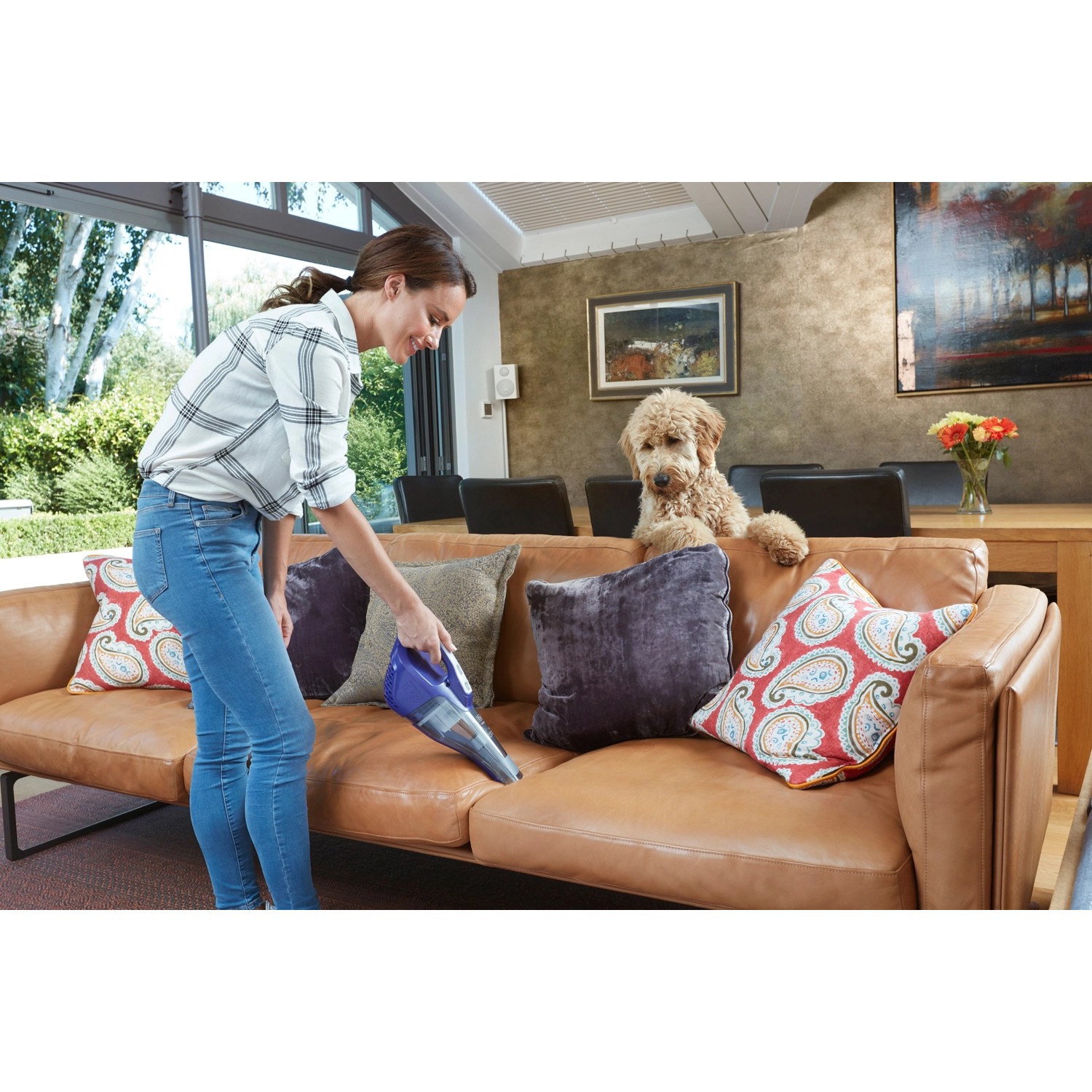 Frau reinigt Sofa mit Black+Decker Akku-Handstaubsauger Pet. Hund sitzt daneben.