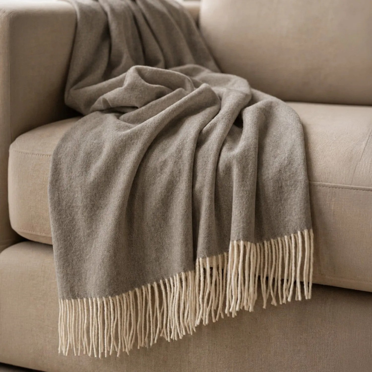 Taupe Baumwoll-Mix Sofadecke mit hellen Fransen, weich auf beigem Sofa drapiert. Leichte Wohndecke 130x170 cm.