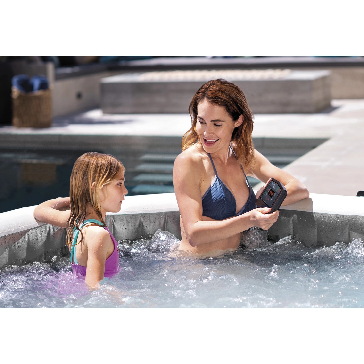 Intex PureSpa Greywood Deluxe: Aufblasbarer Whirlpool mit Mutter und Tochter im Wasser, Steuereinheit in der Hand.
