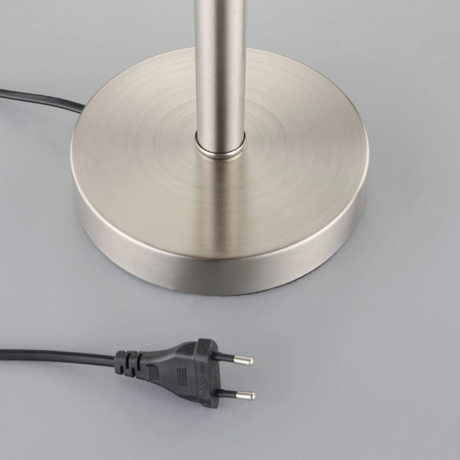 Runde, gebürstete Metallbasis der Lindby Jolie Tischlampe mit schwarzem Eurostecker auf grauem Untergrund.