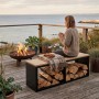 Schwarzes osoltus Brennholzregal mit heller Sitzbank, gefüllt mit Holz, auf Terrasse am Meer mit Person und Feuerstelle.