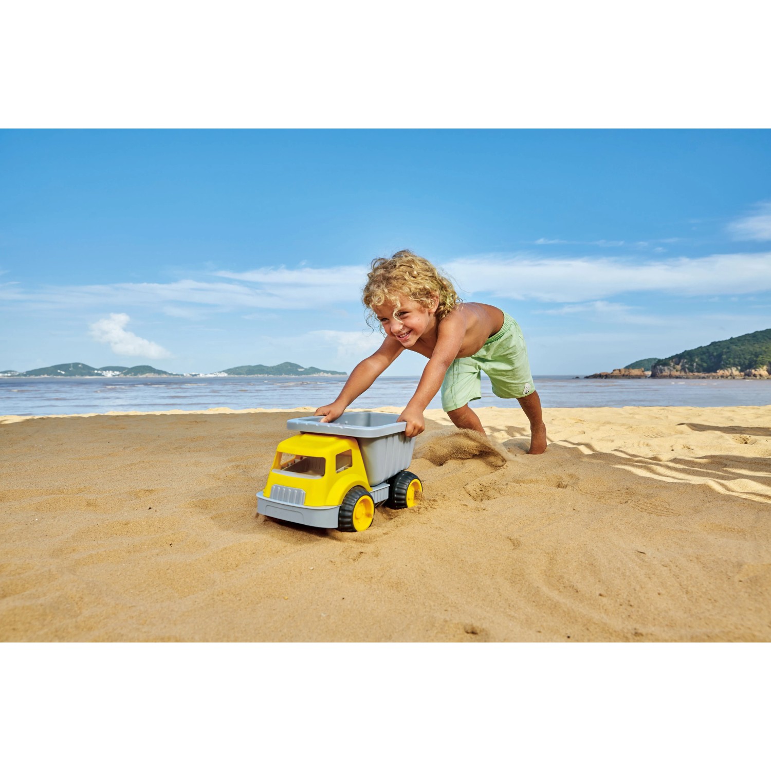Kind spielt mit gelbem Hape Sandlaster am Strand. Robustes Spielzeug für Sandkasten und mehr.