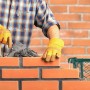 Handwerker in kariertem Hemd und gelben Handschuhen mauert rote Ziegelsteine mit Mörtel und Kelle auf einer Baustelle.