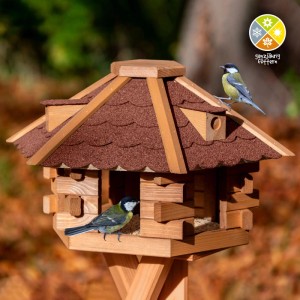 Sechseckiges Dobar Vogelhaus Blockhaus aus Holz mit rotem Bitumendach. Zwei Vögel fressen am Futterhaus.