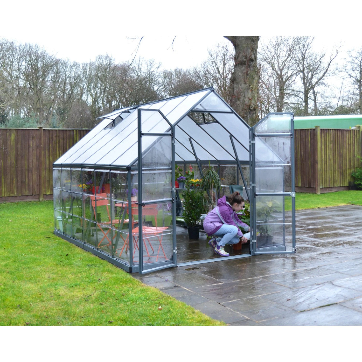 Graues Palram Canopia Gewächshaus 9 m² mit Fundamentrahmen und Person bei der Gartenarbeit.