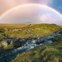 Vlies Fototapete mit Regenbogen über grüner Landschaft und Bachlauf.