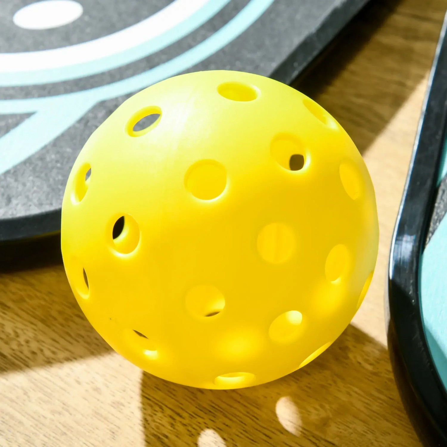 Gelber Pickleball-Ball mit Löchern vor Schläger des SPORTNOW Pappelholz Schläger-Sets.