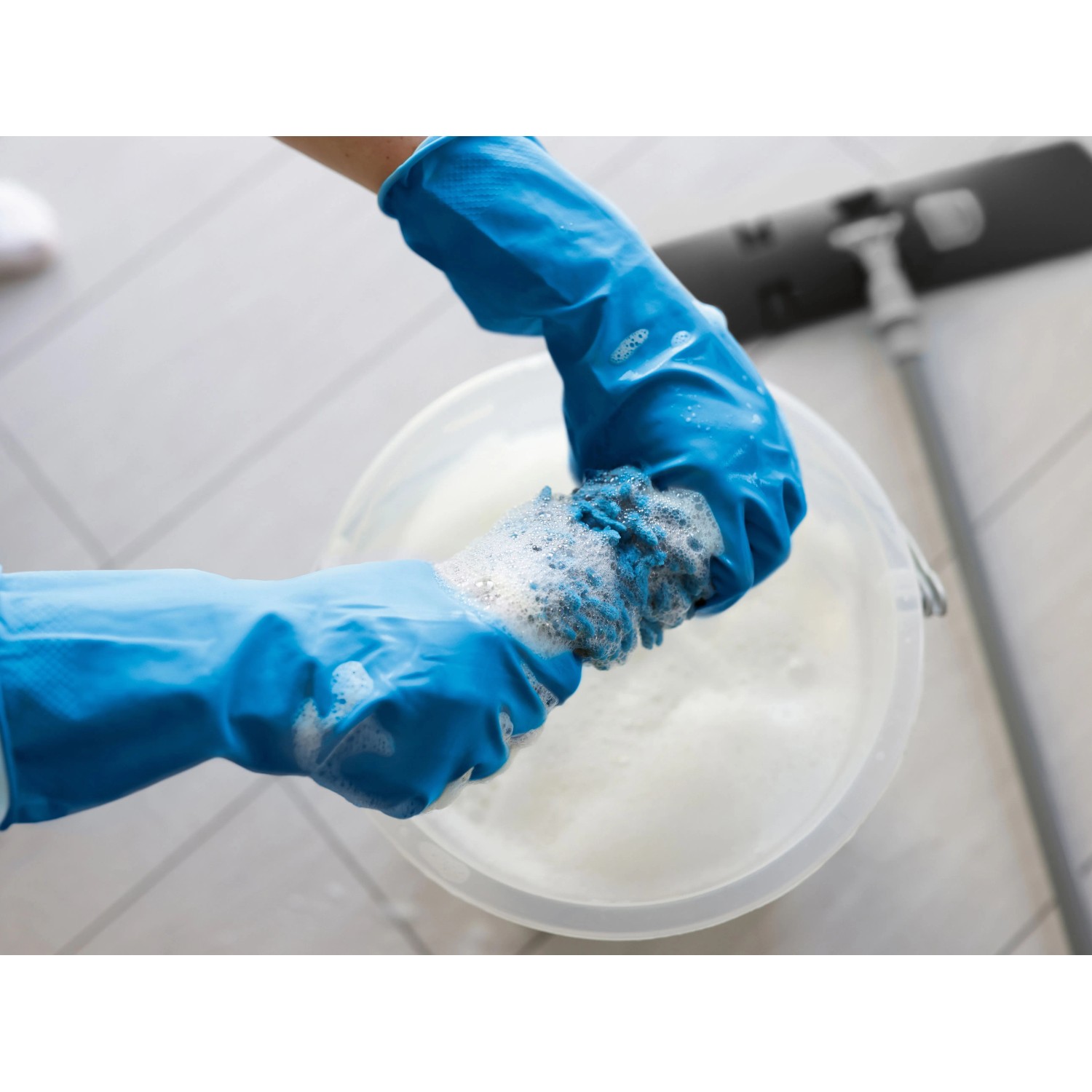 Hände mit blauen Mehrweghandschuhen Robust Größe S wringen einen Schwamm in einem Eimer mit Seifenlauge aus.