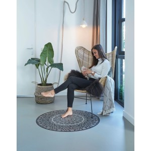 Runder, schwarzer Teppich Smuq Mandela (Ø 100 cm) im Wohnraum mit sitzender Person.