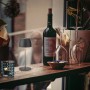 Brilo LED-Akku-Tischleuchte Serina Mini in Anthrazit auf Tisch mit Weinflasche und Gläsern.