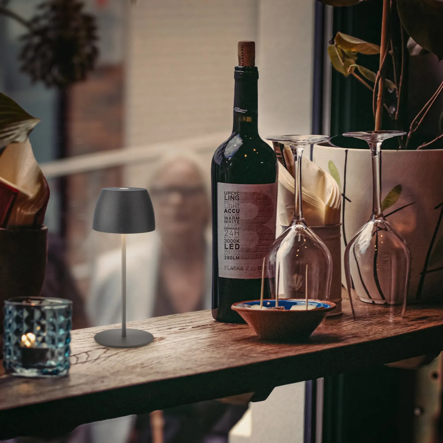 Brilo LED-Akku-Tischleuchte Serina Mini in Anthrazit auf Tisch mit Weinflasche und Gläsern.
