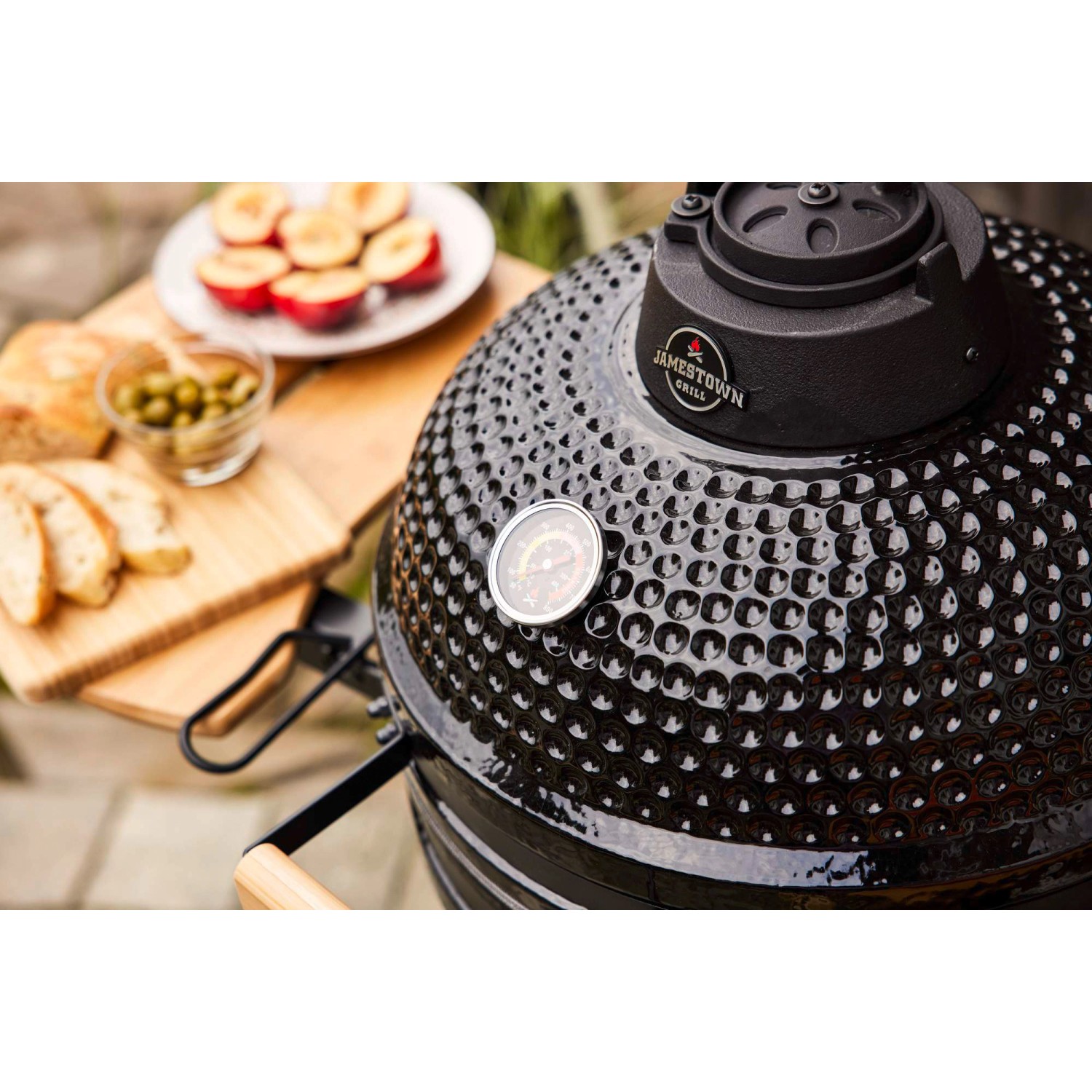 Schwarzer Jamestown Keramik-Holzkohlegrill Marwin M mit Thermometer und Speisen auf einem Tisch.