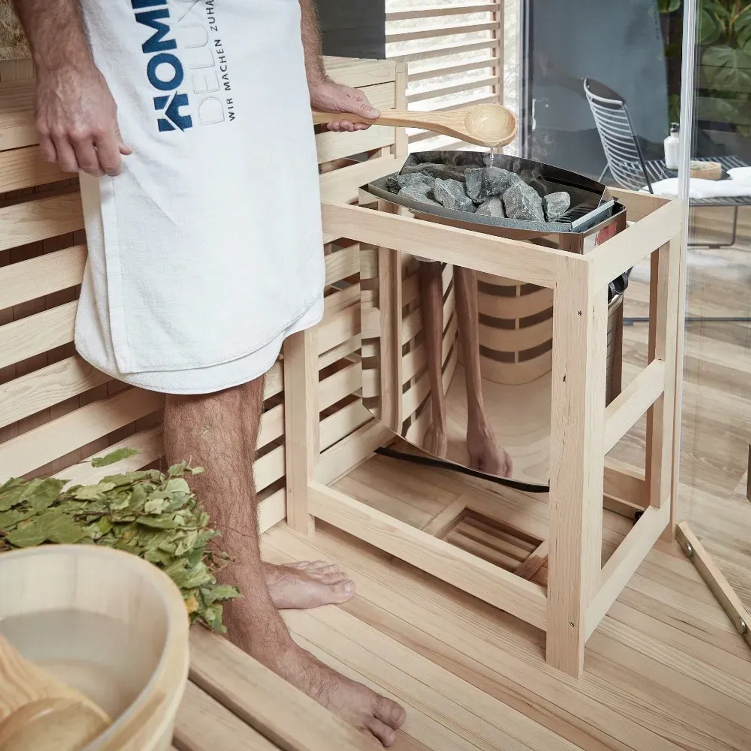 Mann gießt Wasser auf Steine im Saunaofen der SKYLINE BIG Tannenholz Sauna. Zubehör und Handtuch sind sichtbar.