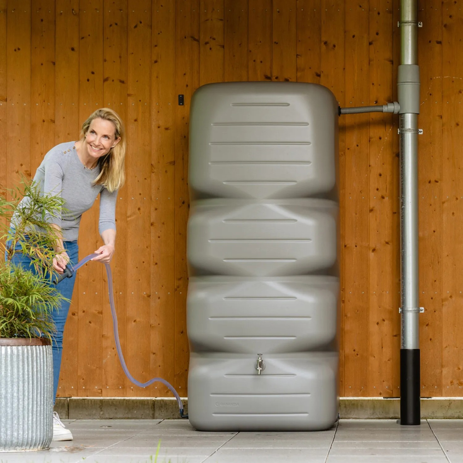 Garantia Gartentank Cubus 700 l in Betongrau mit Frau und Schlauch zur Gartenbewässerung.