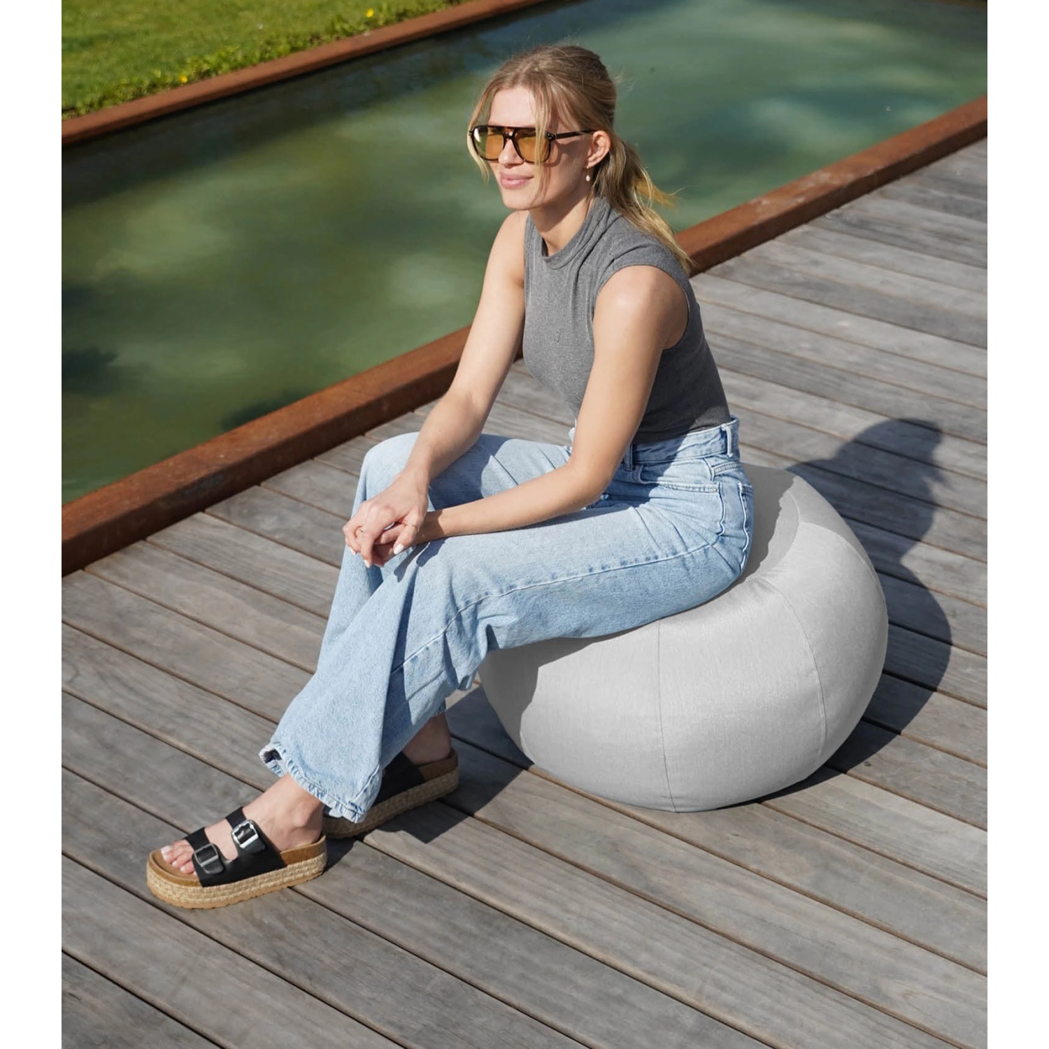 Frau sitzt auf mokebo Outdoor Pouf 'Der Kugelige' in Hellgrau, ideal als Sitzhocker oder Bodenkissen für Balkon und Garten.