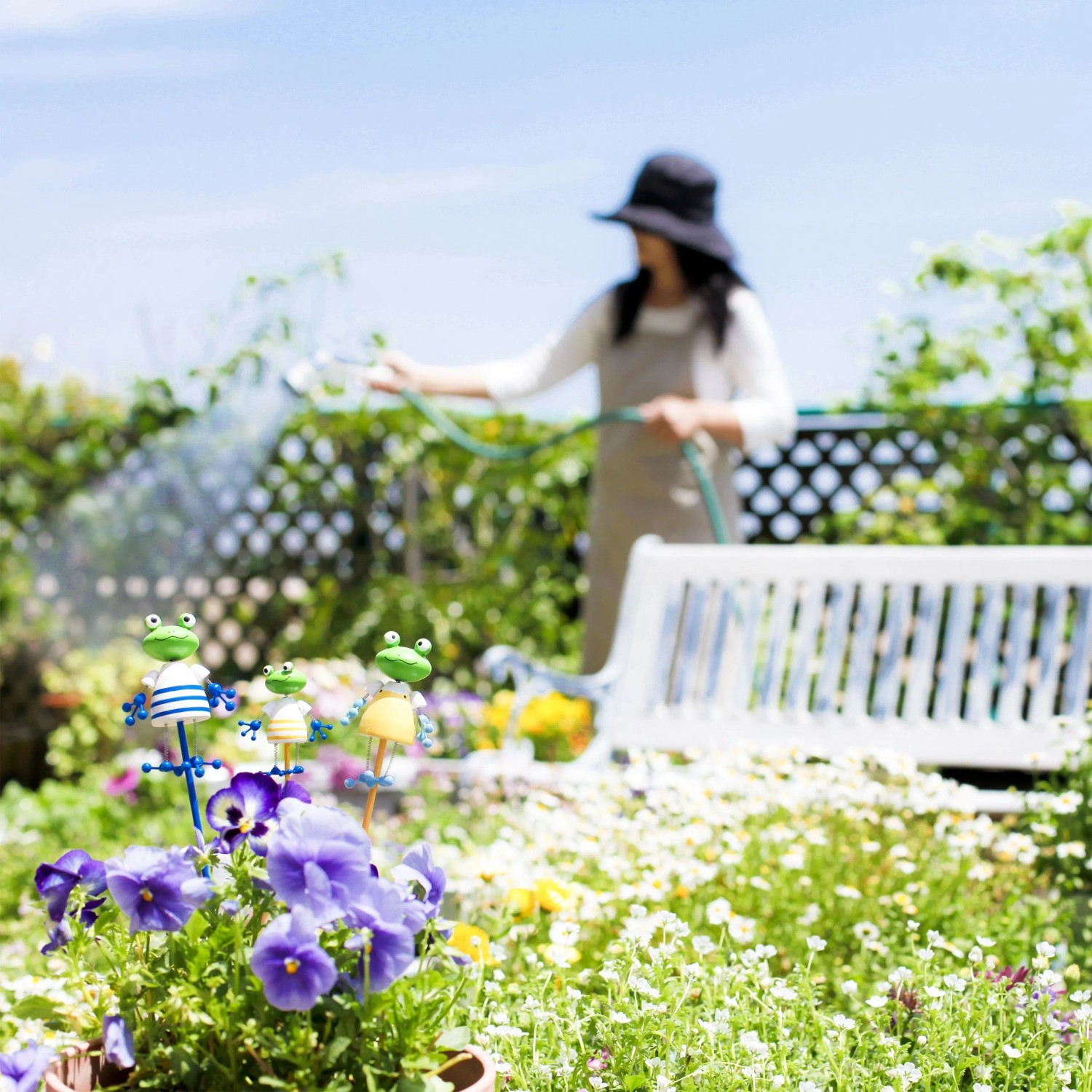 Person gießt Blumen im Garten mit Schlauch; im Vordergrund lila Blüten und Froschfiguren.