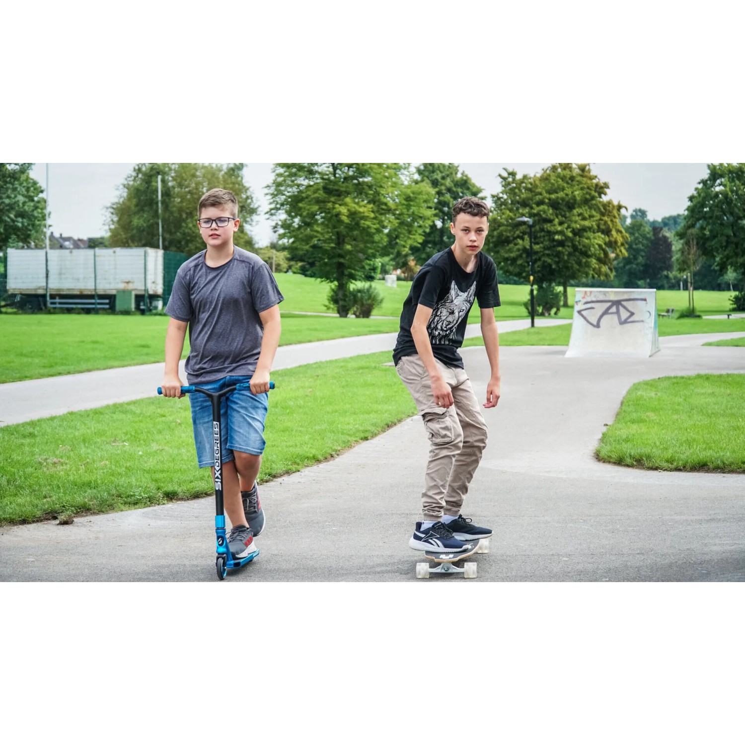 Zwei Jugendliche mit blauem Six Degrees Stuntscooter und Skateboard im Skatepark.