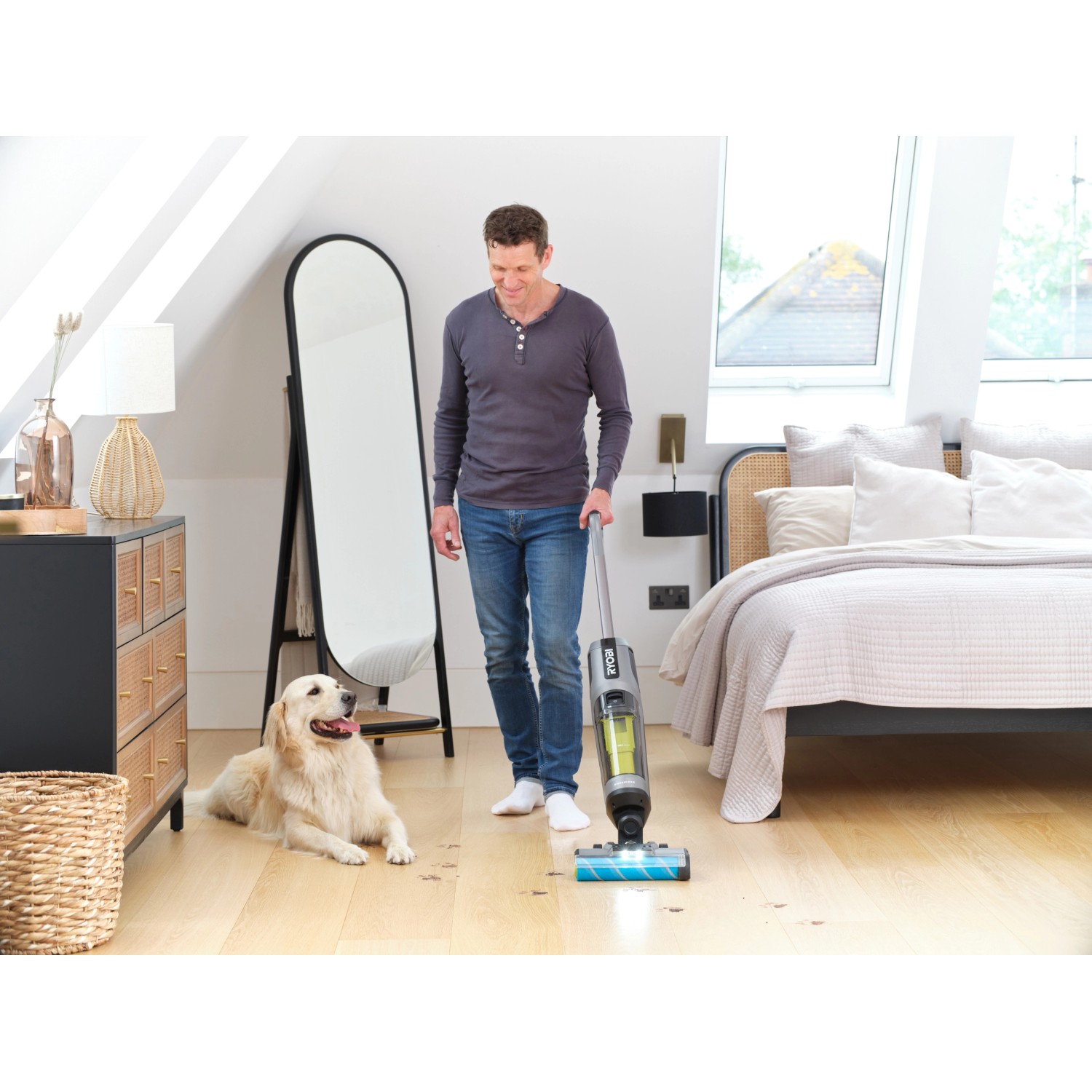 Mann reinigt Holzboden mit Ryobi Multiflächenreiniger. Hund liegt daneben.