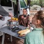 Petromax Atago Gasgrill-Set beim Camping: Grill, Mais und Personen am Tisch im Freien.