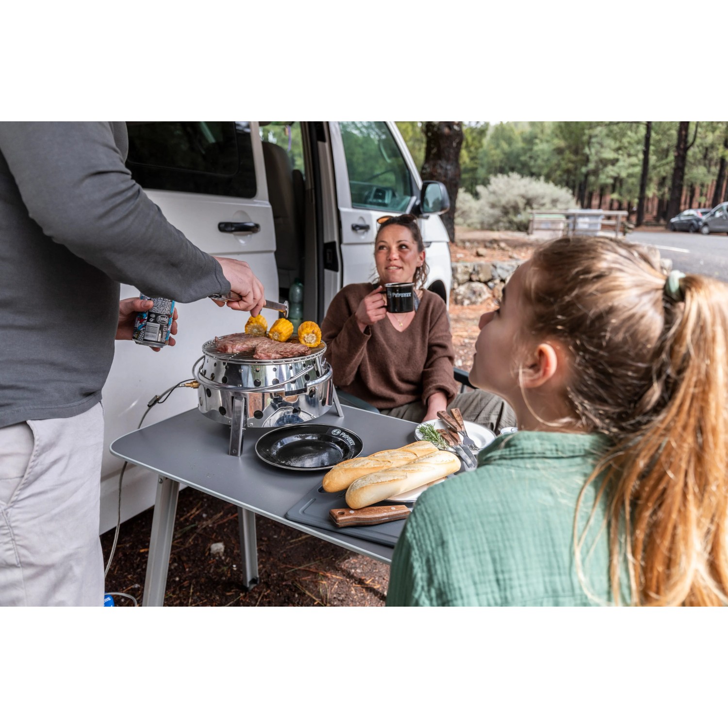 Petromax Atago Gasgrill-Set beim Camping: Grill, Mais und Personen am Tisch im Freien.