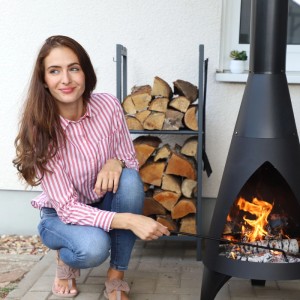 Frau schürt Feuer in Ofen, daneben anthrazitfarbenes Relaxdays Kaminholzregal aus Stahl mit 2 Ablagen voller Holzscheite.