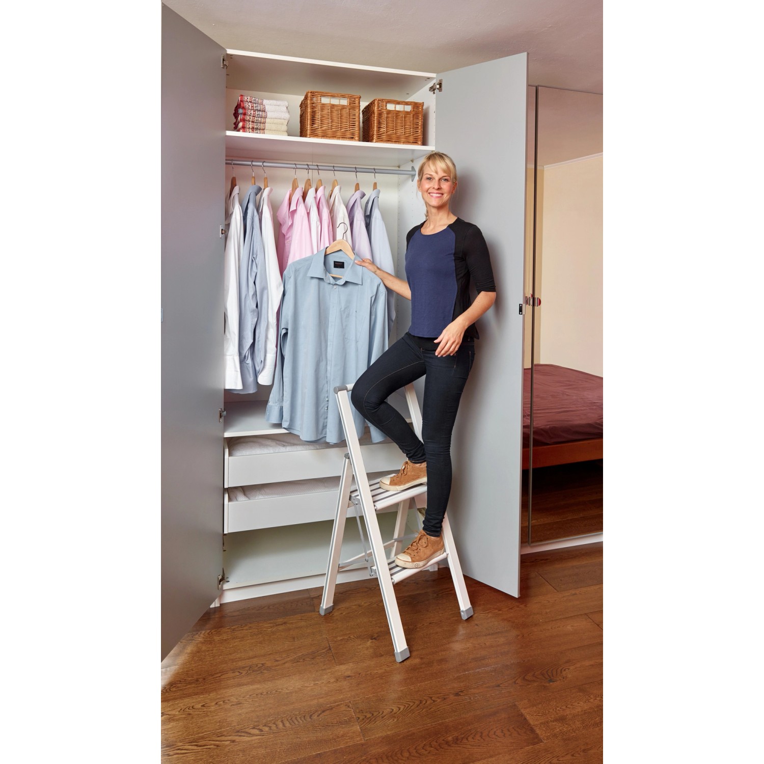 Frau auf Wenko Alu-Klapptritt (2-stufig, weiß) vor Kleiderschrank. Sicheres Erreichen höherer Ebenen.