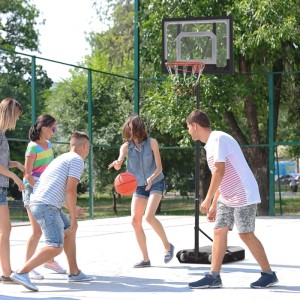 Jugendliche spielen Basketball mit höhenverstellbarem HOMCOM Basketballständer auf Outdoor-Platz. Schwarze Basis, transparentes Backboard.