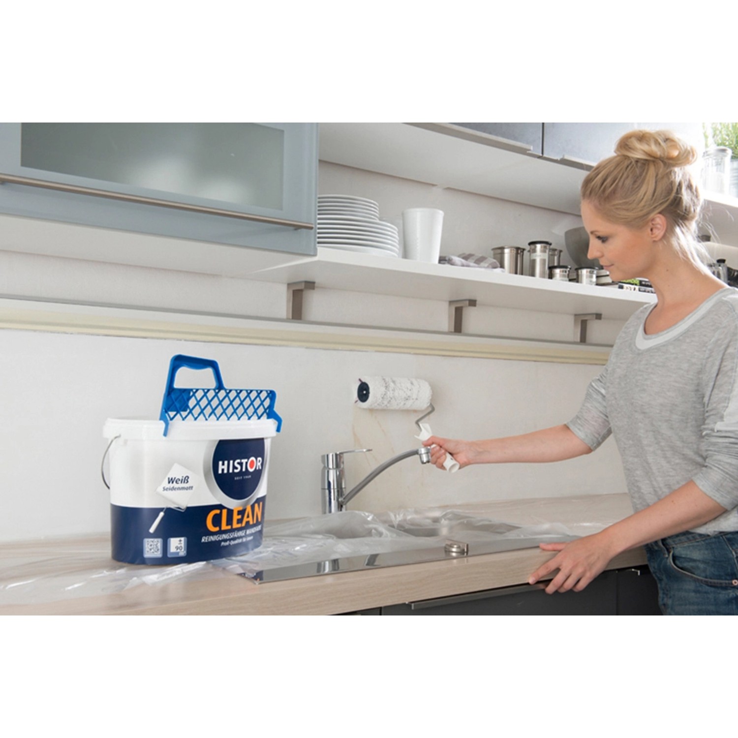 Frau streicht Wand mit Histor Clean Wandfarbe Weiß seidenmatt. 10L Eimer und Farbroller sichtbar.