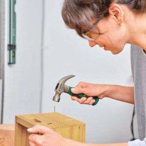 Frau schlägt Nagel mit Bosch Klauenhammer 8oz in Holzkiste. Ergonomischer Griff für präzise DIY-Arbeiten.