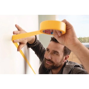 Mann klebt gelbes tesa® Malerband Perfect+ an eine weiße Wand für präzise Farbkanten.