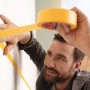 Mann klebt gelbes tesa® Malerband Perfect+ an eine weiße Wand für präzise Farbkanten.