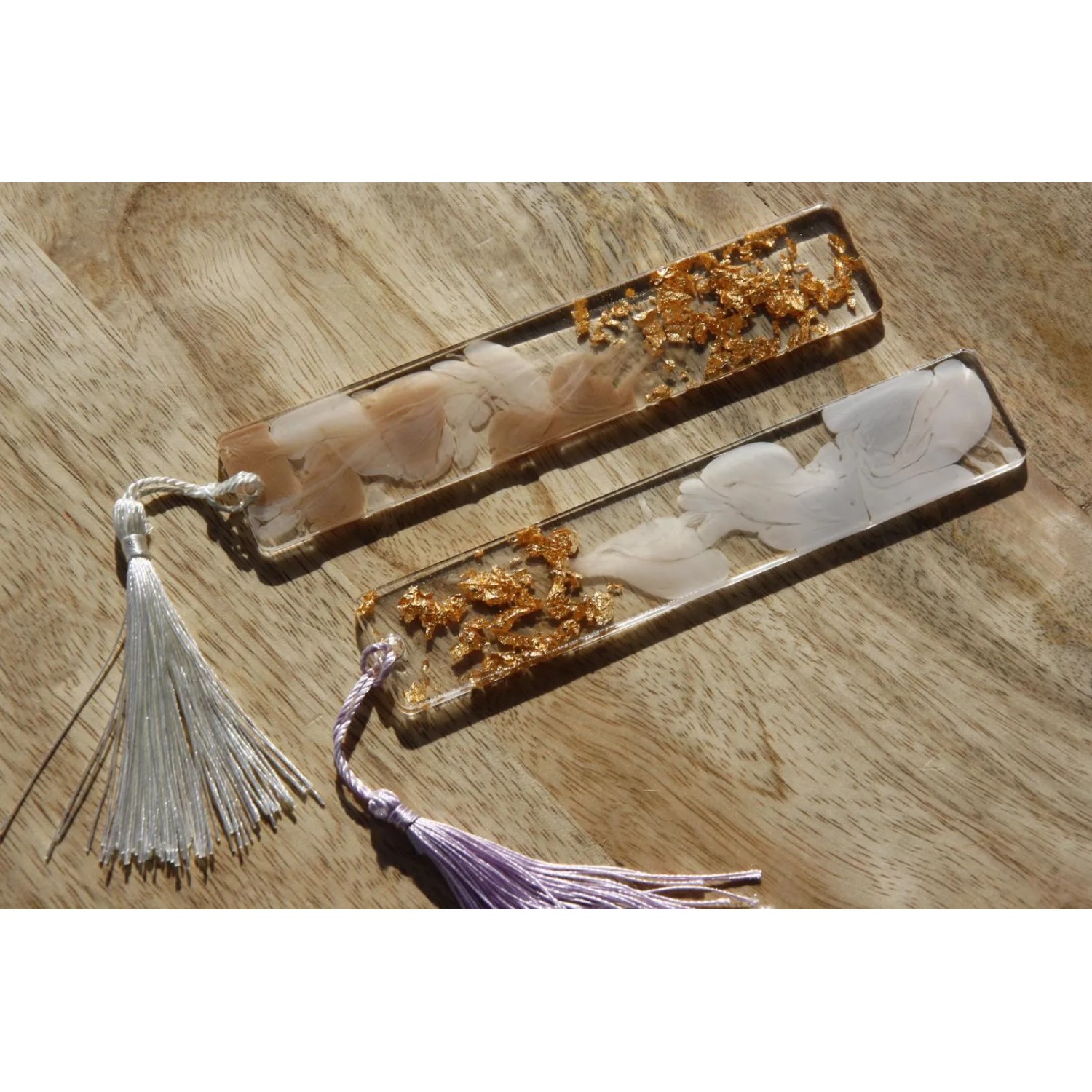 Zwei CreativEpoxy Epoxidharz Lesezeichen mit Quasten und Goldflocken auf Holz.