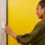 Frau bedient weißen Yale Panikknopf/Türklingel Smart Alarm an Wand neben gelber Tür.