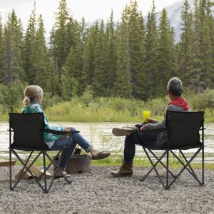 Zwei Personen sitzen auf schwarzen Sunula Campingstühlen mit Getränkehaltern am Flussufer.