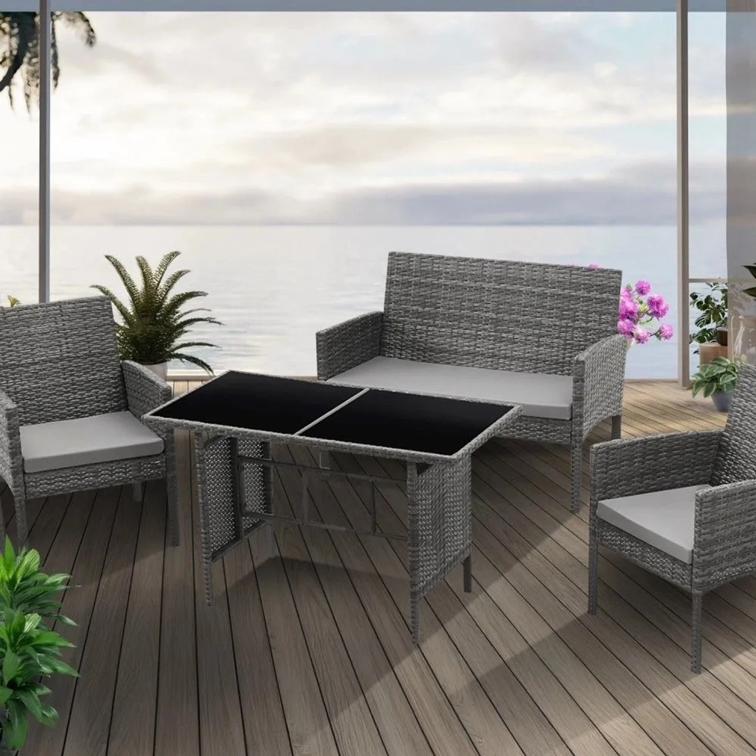 Graue Svita Brooklyn XL Rattan Essgruppe: Tisch mit Glasplatte, 2 Sessel, 2er Sofa mit Kissen auf Holzterrasse.
