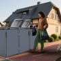 Anthrazitfarbene FM-SOLAR Mülltonnenbox für 3 Tonnen im Garten. Frau entsorgt Abfall, Deckel mit Gasdruckfeder offen.